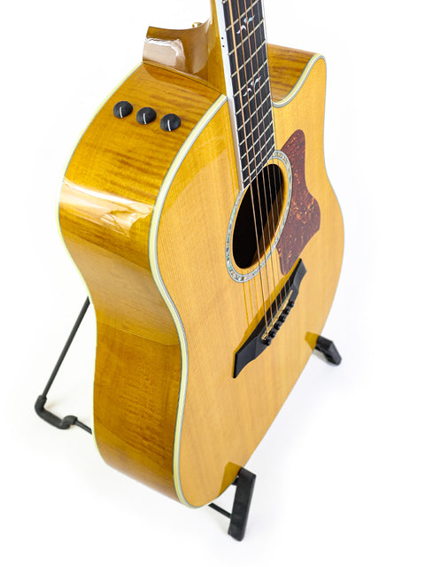 A 2003 Taylor 610CE guitar resting on a stand, set against a bright white backdrop.