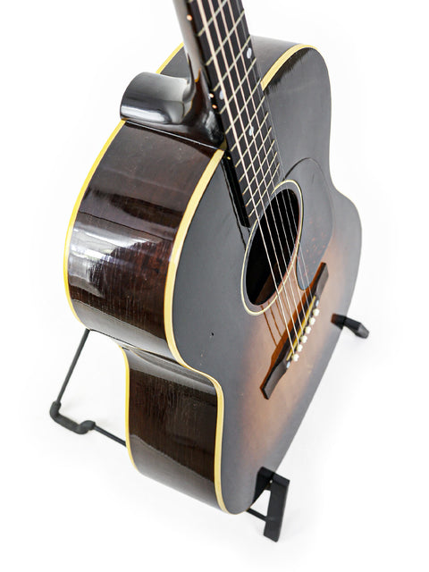 A black Gibson LG-1 acoustic guitar from 1954 displayed on a stand, highlighting its vintage charm and craftsmanship.