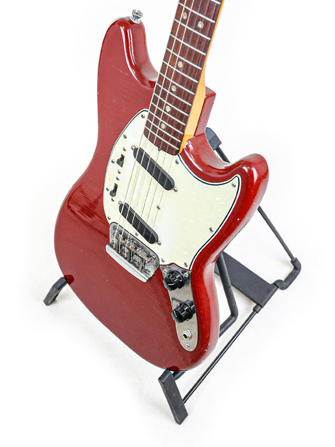 A vibrant red 1966 Fender Musicmaster II guitar resting on a stand, highlighted by a simple white background.