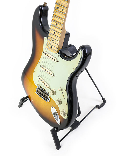Fender John Cruz Master Built ‘64 Stratocaster Relic guitar resting on a stand, set against a bright white background.