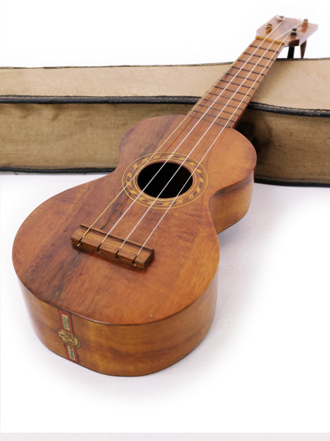 A classic Kumalae Style 2 ukulele, dating back to 1913, is displayed in a wooden case next to a book.