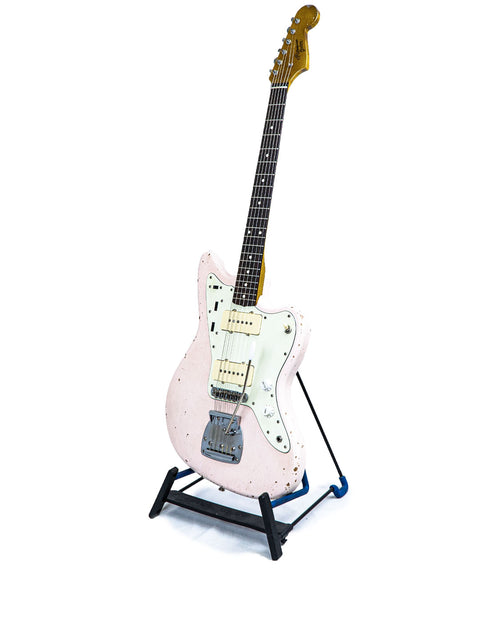 A pink guitar, model Rittenhouse JM-63 from 2020, elegantly positioned on a stand with a simple white backdrop.