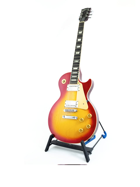 A white background highlights a Gibson Les Paul Standard guitar from 1998, elegantly positioned on a stand.