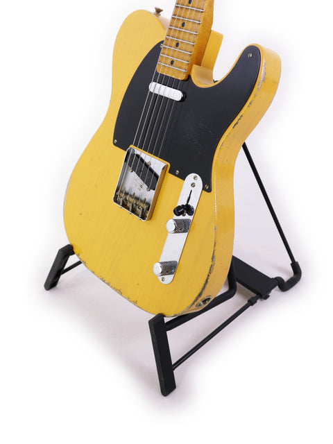 A yellow guitar on a stand, the Fender Carlos Lopez Masterbuilt Nocaster from 2019, adorned with a black strap.