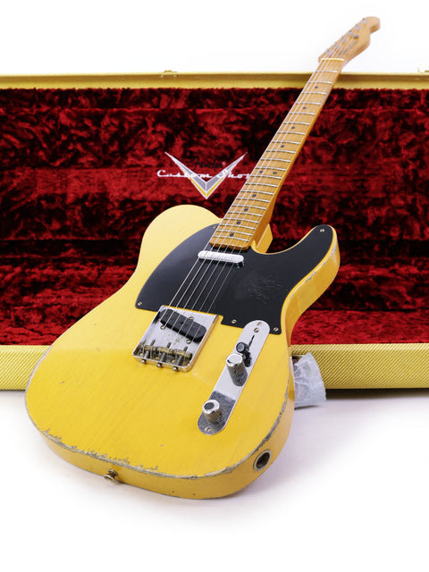 A yellow guitar, the Fender Carlos Lopez Masterbuilt Nocaster, rests in a case next to a red box, highlighting its unique design.