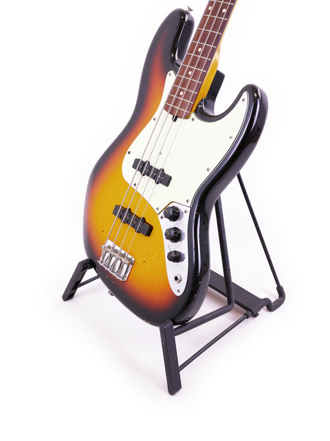Fender Custom Shop '64 Jazz Bass Relic displayed on a stand with a minimalist white backdrop.