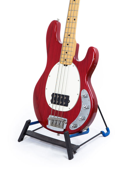 Red Music Man Stingray 4 H bass guitar displayed on a stand, with a blue foot pedal nearby.