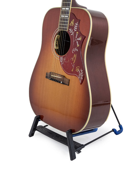 The image features a Gibson Hummingbird True Vintage guitar with a rich brown body, elegantly supported by a black stand.