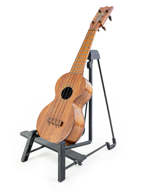 1920s Kumalae ukulele sits on a stand, highlighting its vintage charm and Hawaiian heritage in a cozy setting.