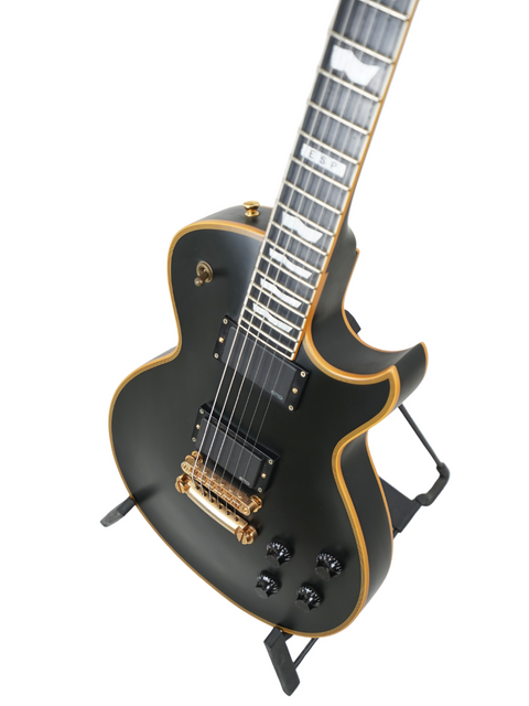 A black electric guitar with gold trim and details displayed on a stand against a white background.