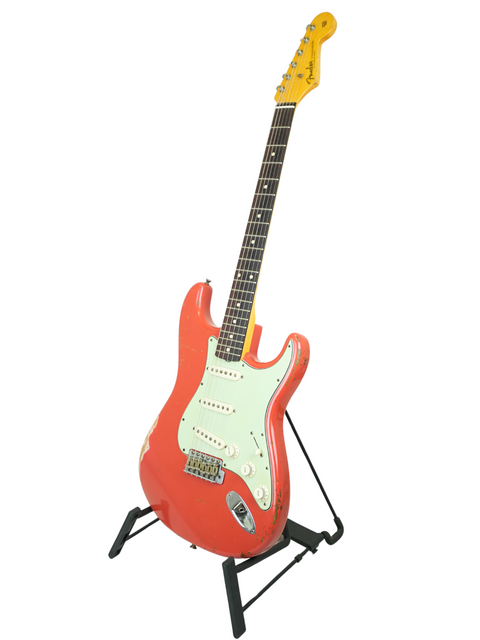 A red Fender Kyle McMillin Masterbuilt 1960 Stratocaster Relic guitar on a stand against a white background.
