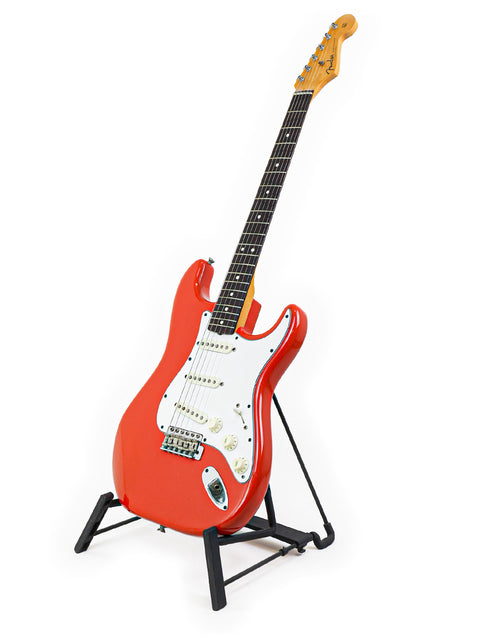 A striking red Fender AVRI ‘62 Stratocaster guitar resting on a stand, set against a simple white background.
