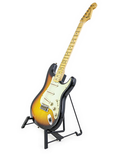 Displayed on a stand, a Fender John Cruz Master Built ‘64 Stratocaster Relic guitar contrasts with a white backdrop.