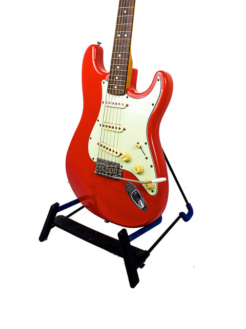 A striking red Fender Strat guitar, model 62 AVRI, displayed on a black stand, highlighting its classic Fiesta Red color.
