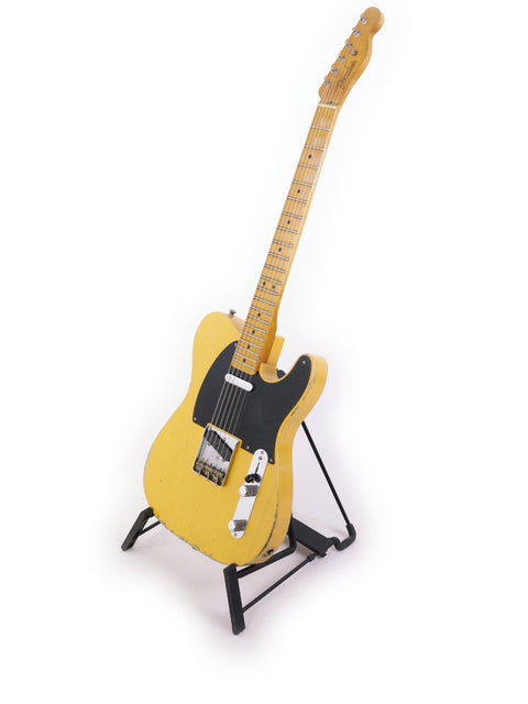A striking yellow Fender Carlos Lopez Masterbuilt Nocaster guitar on a stand, highlighted by a simple white background.