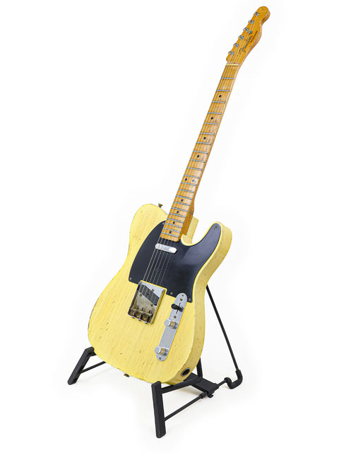 A striking yellow electric guitar rests on a stand, representing the Fender Master Built Carlos Lopez 70th Anniversary Broadcaster, USA 2020.