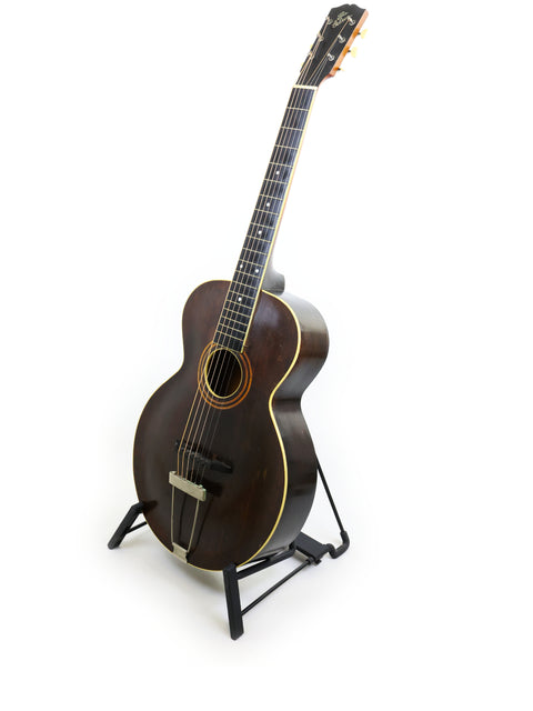 A vintage 1920 Gibson L-1 guitar on a stand, highlighted by a stark white background.