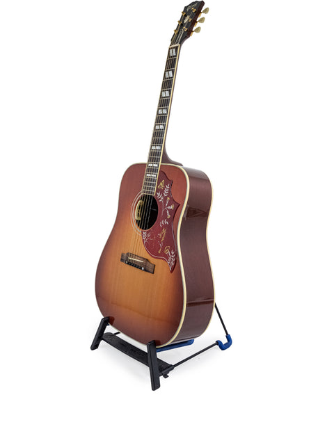 Gibson Hummingbird True Vintage guitar on a stand against a clean white background, showcasing its elegant design.
